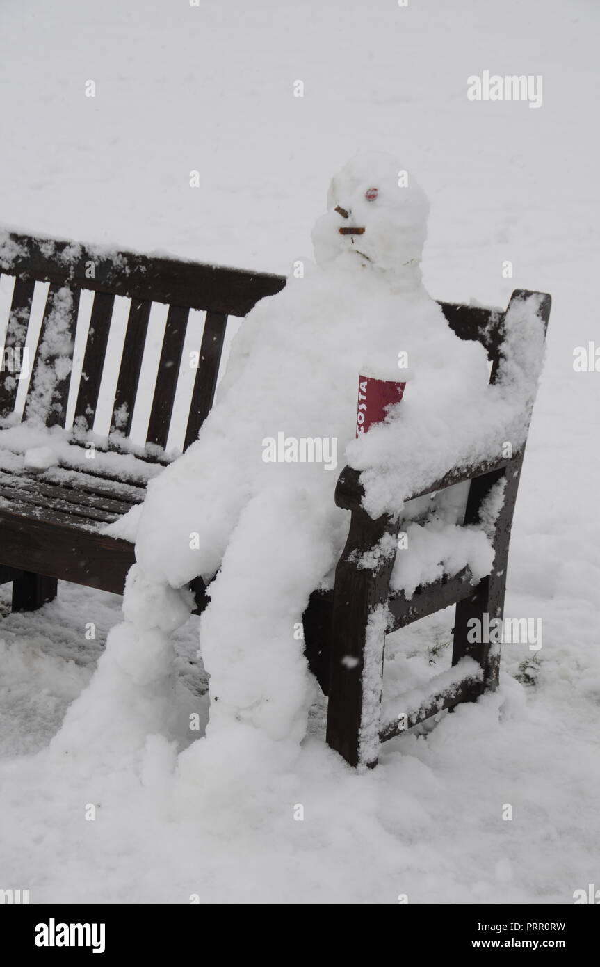 Ein sitzender Schneemann entspannen auf einer Parkbank mit seiner warmen Tasse Kaffee während einer Snow Fall in Marlow, Buckinghamshire, Großbritannien Stockfoto