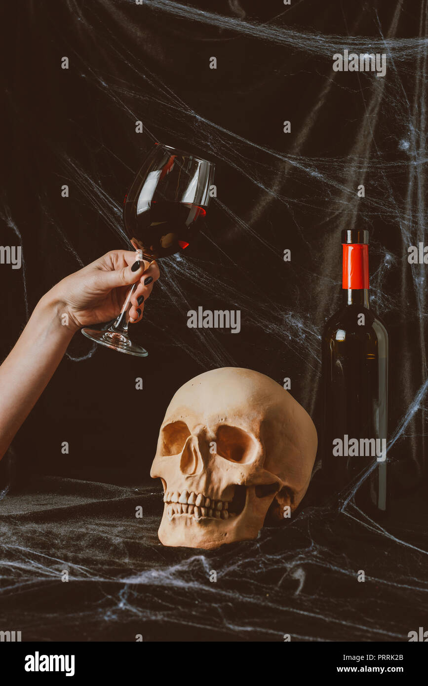 7/8-Ansicht von Frau mit Glas Rotwein in der Nähe von Totenkopf auf schwarzem Stoff mit Spinnennetz Stockfoto