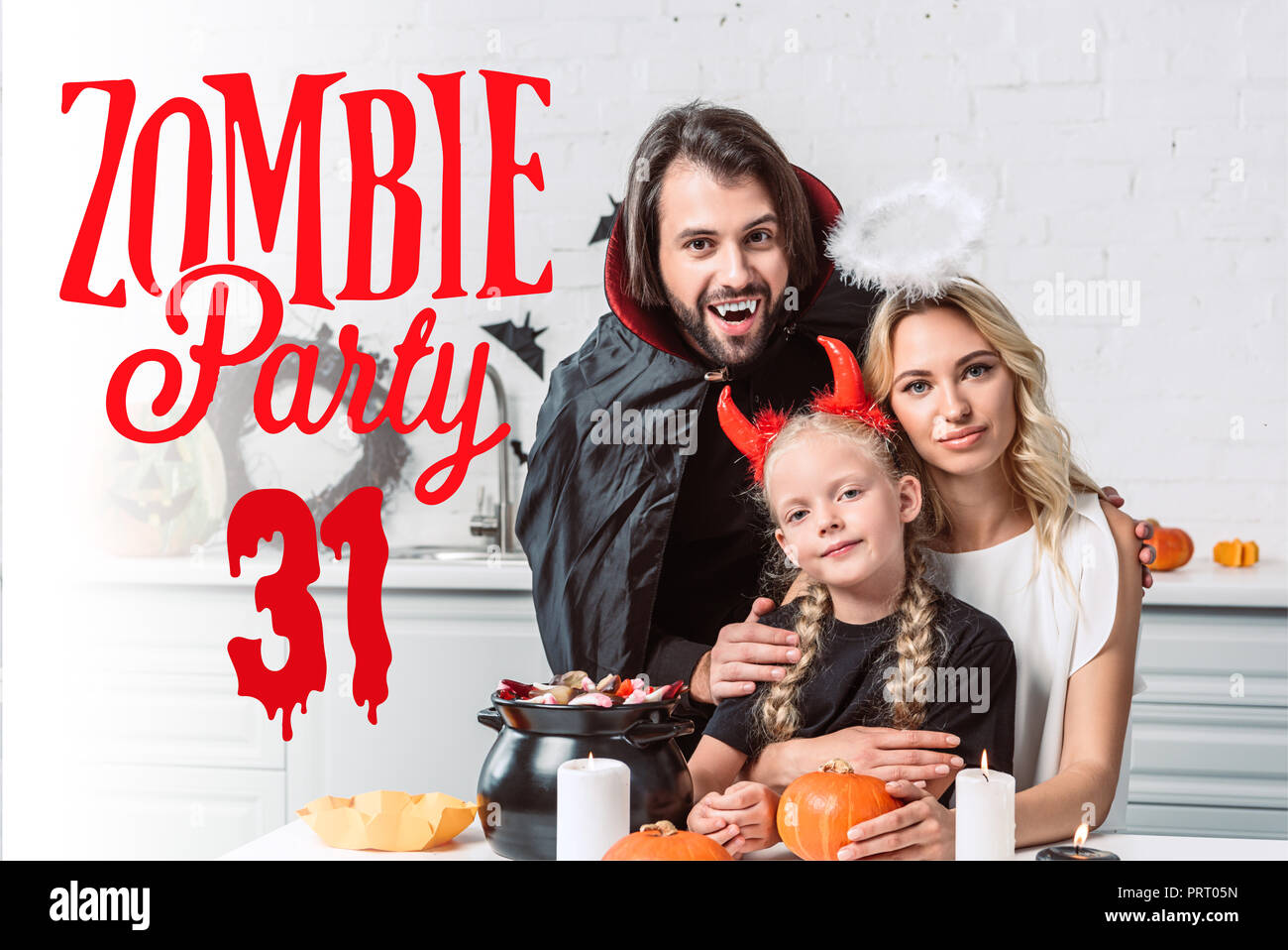 Portrait von Eltern und Tochter in Halloween Kostüme bei Tisch mit Leckereien im schwarzen Topf in der Küche zu Hause mit 'Zombie party 31'-Schriftzug Stockfoto
