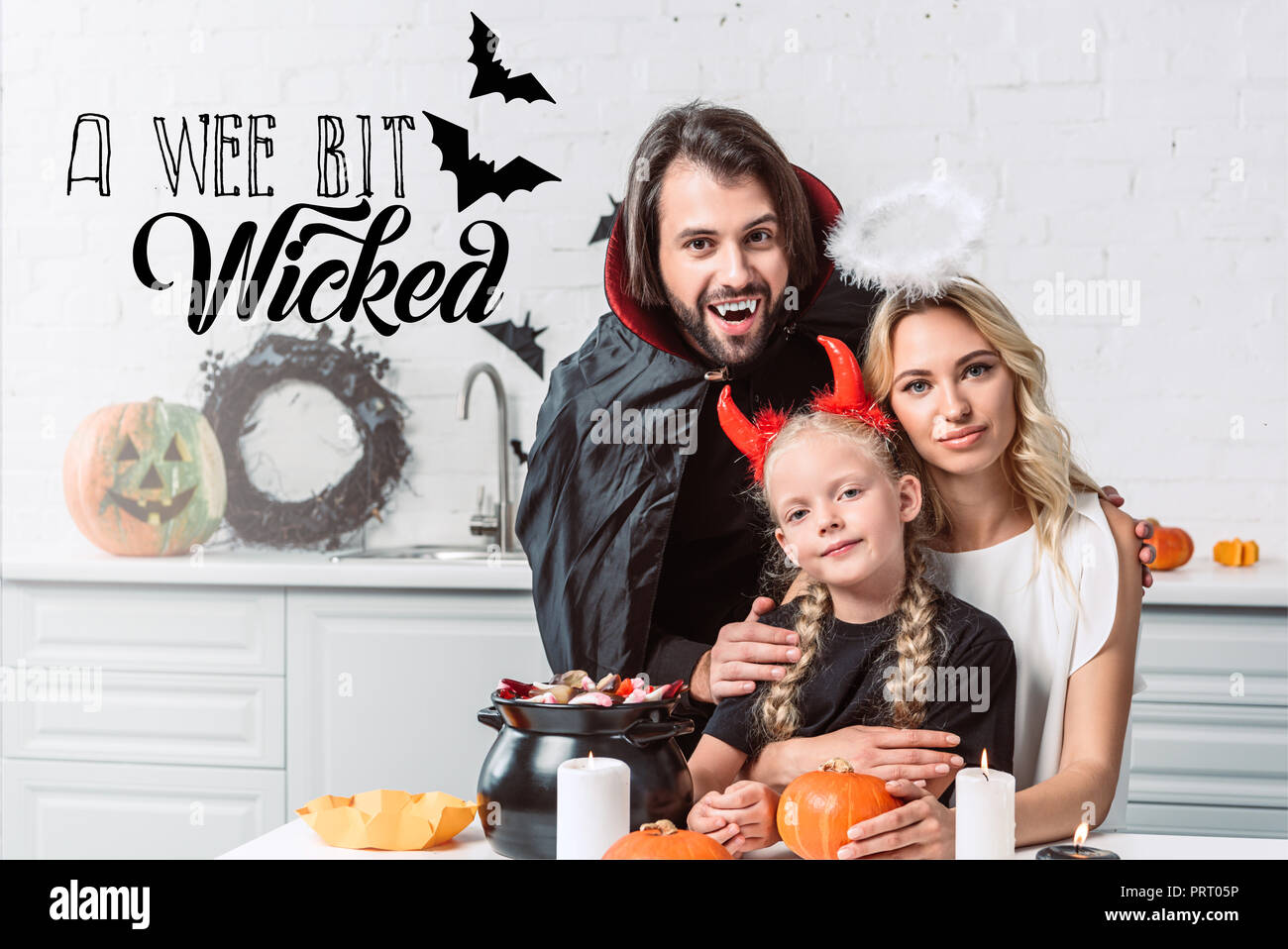 Portrait von Eltern und Tochter in Halloween Kostüme bei Tisch mit Leckereien im schwarzen Topf in der Küche zu Hause mit 'ein bisschen böse' Schriftzug Stockfoto