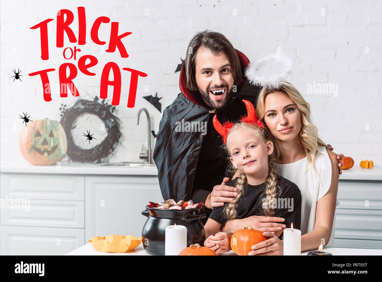 Portrait von Eltern und Tochter in Halloween Kostüme bei Tisch mit Leckereien im schwarzen Topf in der Küche zu Hause mit 'Trick or Treat' Schriftzug Stockfoto