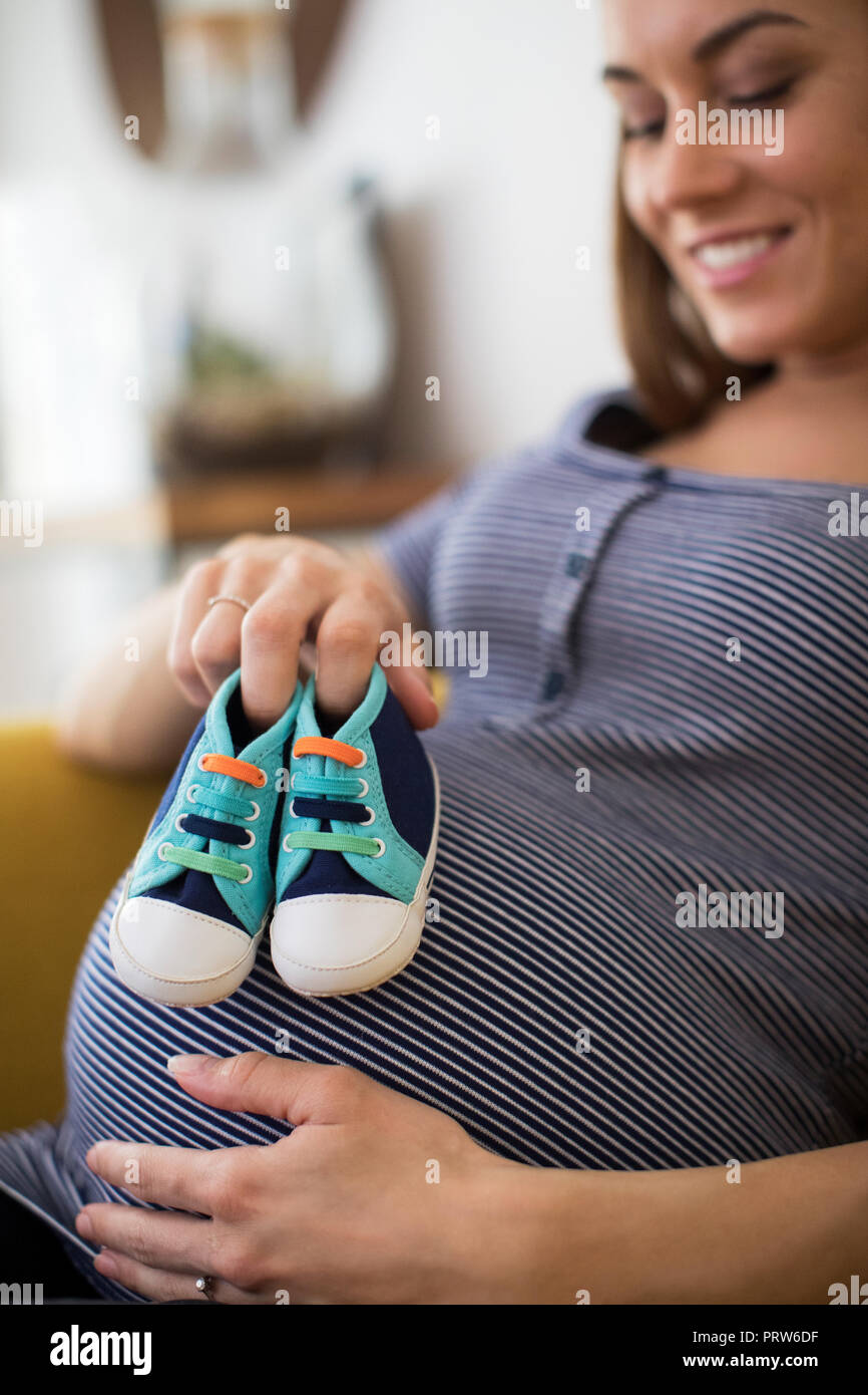 Schwangere Frau Sittingh auf dem Sofa zu Hause Holding Baby Schuhe Stockfoto