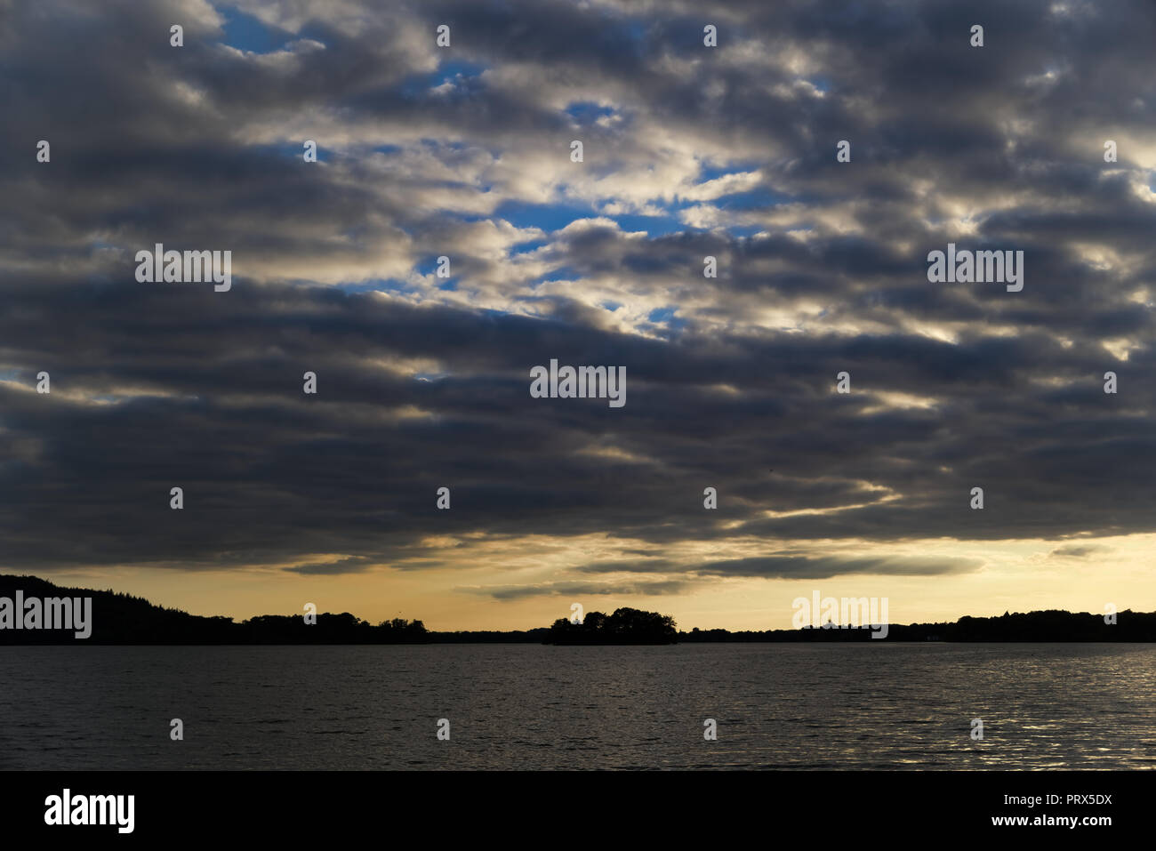 Dramatische Himmel bei Sonnenuntergang über dem See mit Wald am Horizont Stockfoto