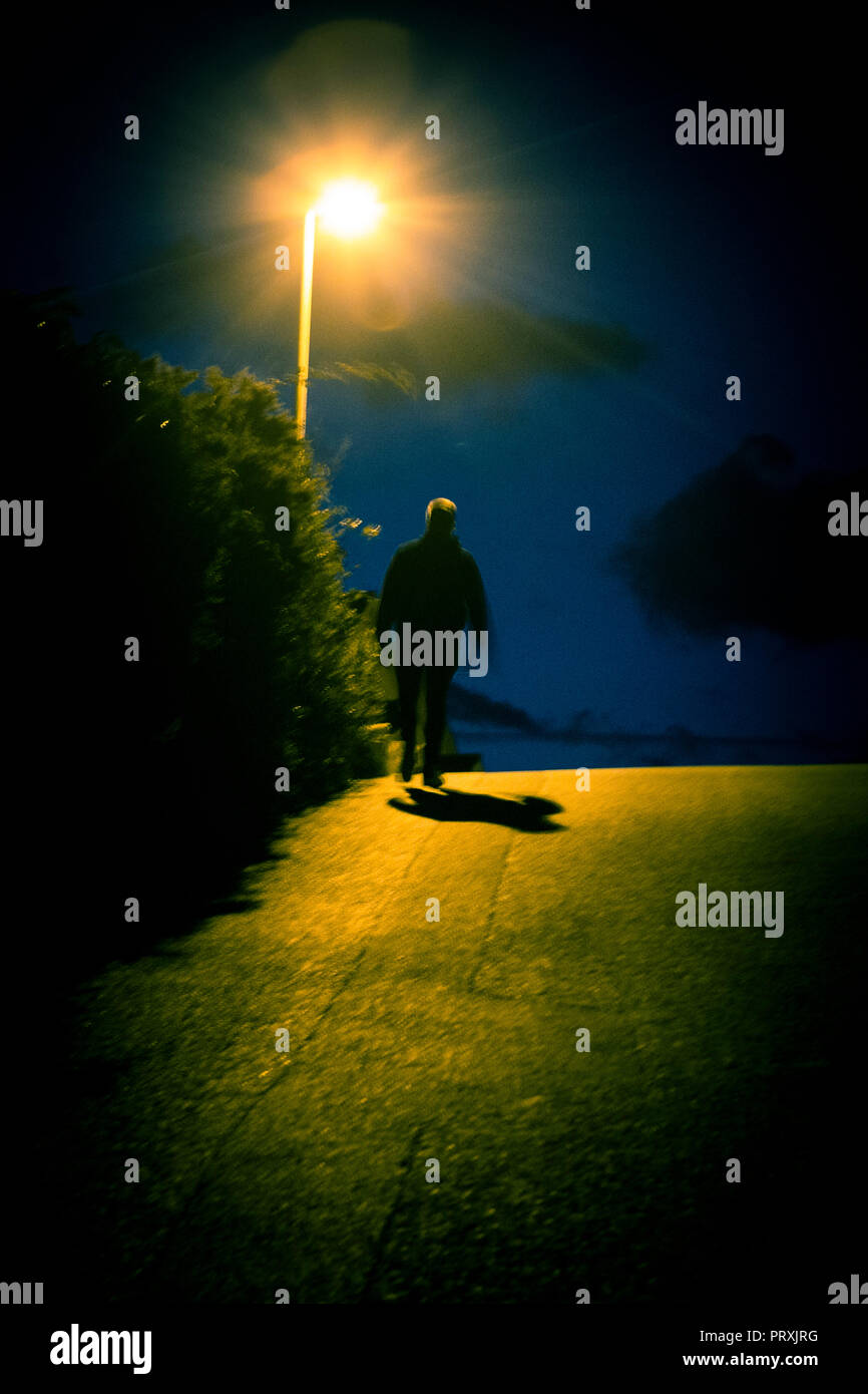 Eine vermummte Gestalt geht auf einen Hügel in der Nacht in silhoutte von der Straßenlaterne casting Licht gesehen, die auf den umliegenden Straßen. Stockfoto