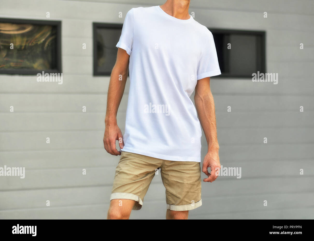 Städtische Vorlage von Kleidung. Yong Mann in eine leere T-Shirt und Shorts Braun stand in der Nähe der weißen texturierte Wand mit schwarzen Fenstern. Mockup kann für y verwenden Stockfoto