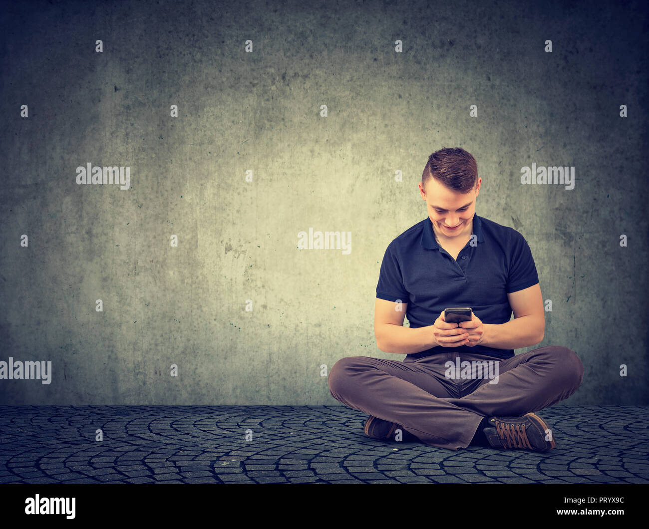 Junger Mann mit Handy und Chatten mit Lächeln saß auf dem Boden vor grauem Hintergrund Stockfoto