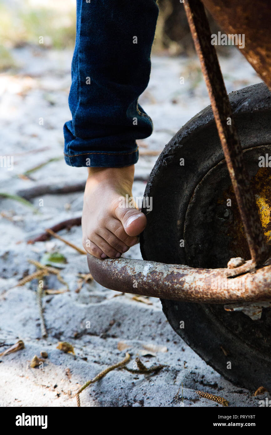 Childs rechten Fuß stehend barfuß auf dem Stahl, bei der eine rostige Rad barrel. Kind jeanswearing tragen Jeans. Stockfoto