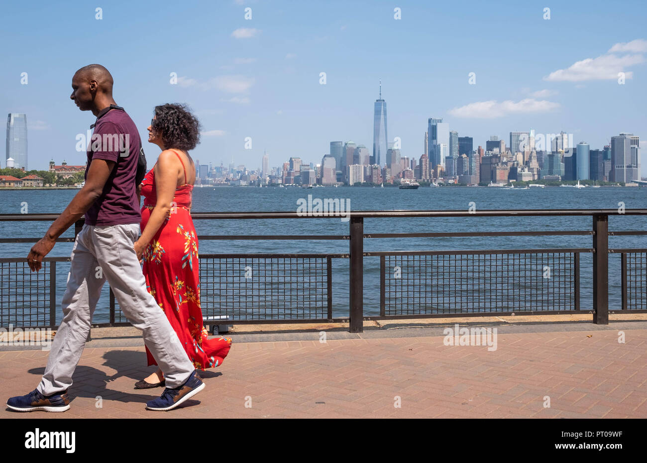 Ein paar gehen Hand in Hand auf Liberty Island mit der New York City Skyline im Hintergrund, einschließlich Lower Manhattan und der Freedom Tower Stockfoto