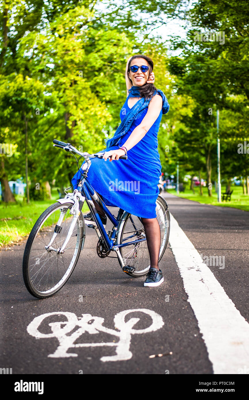 Junge brünette Mädchen im blauen Kleid und Sonnenbrille sitzt auf einem Fahrrad auf einem Fahrrad auf asfalt Straße. Stockfoto