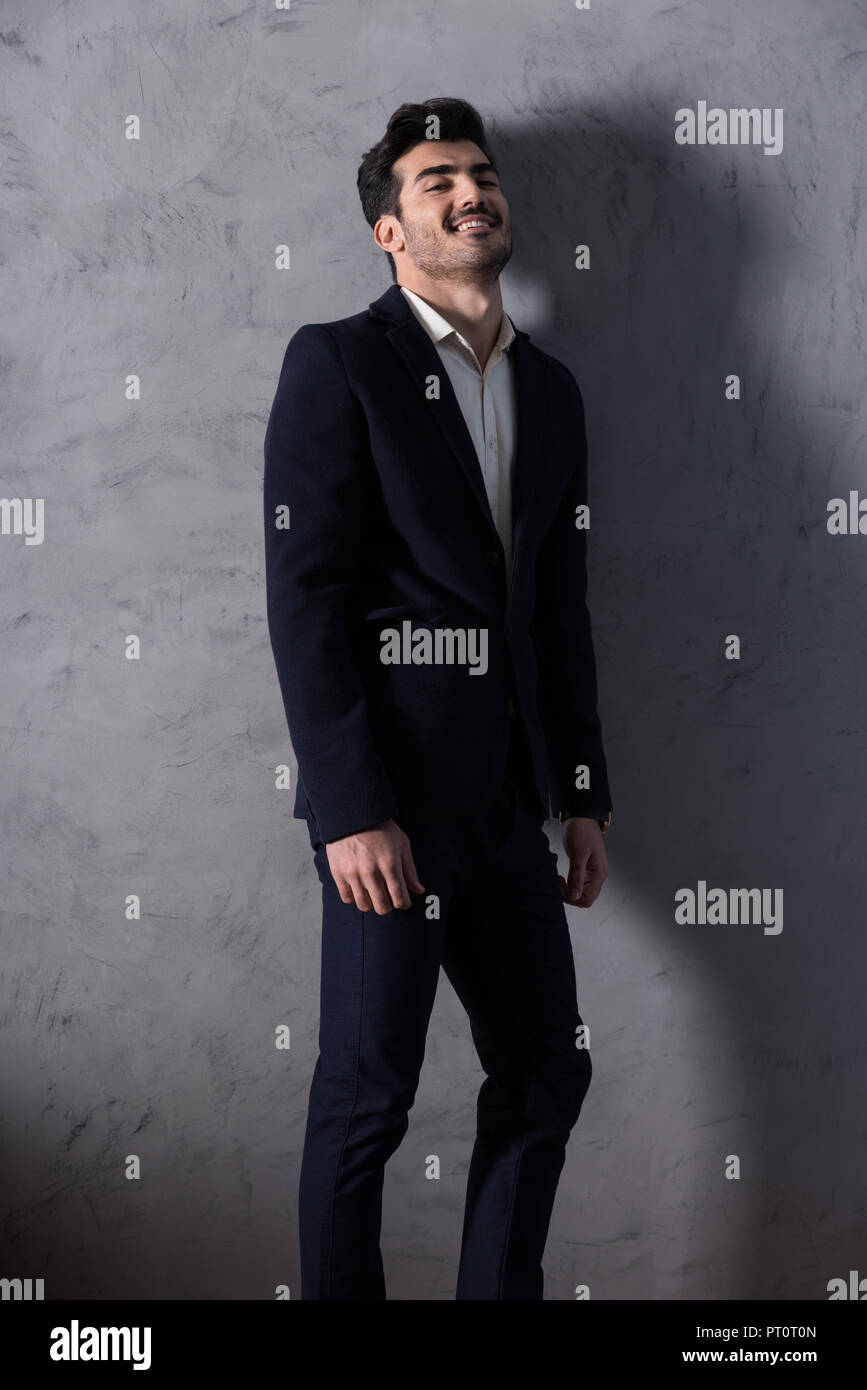 Ein glücklicher hübscher junger Geschäftsmann in einem schwarzen Anzug stand vor einer grauen Wand in einem Studio. Stockfoto