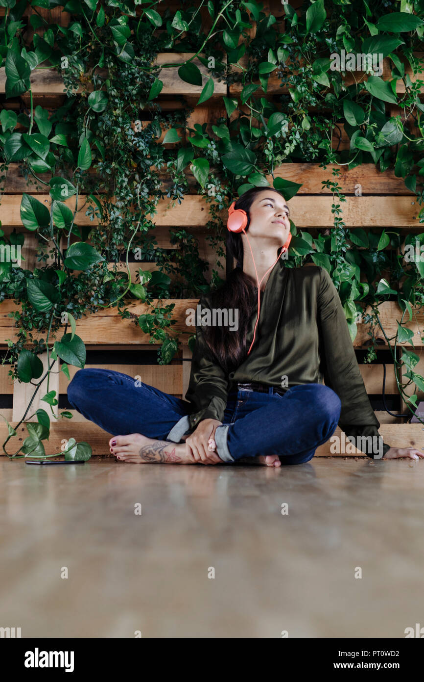 Junge Frau sitzt auf dem Boden an der Wand mit kletterpflanzen Musik hören Stockfoto