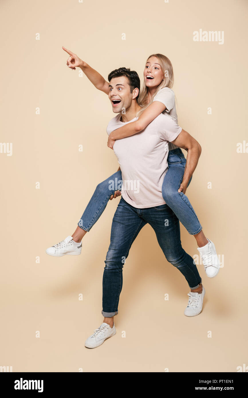 In voller Länge Porträt einer fröhlichen jungen Paar ständigen zusammen über beigen Hintergrund, piggyback Ride, weg zeigend Stockfoto