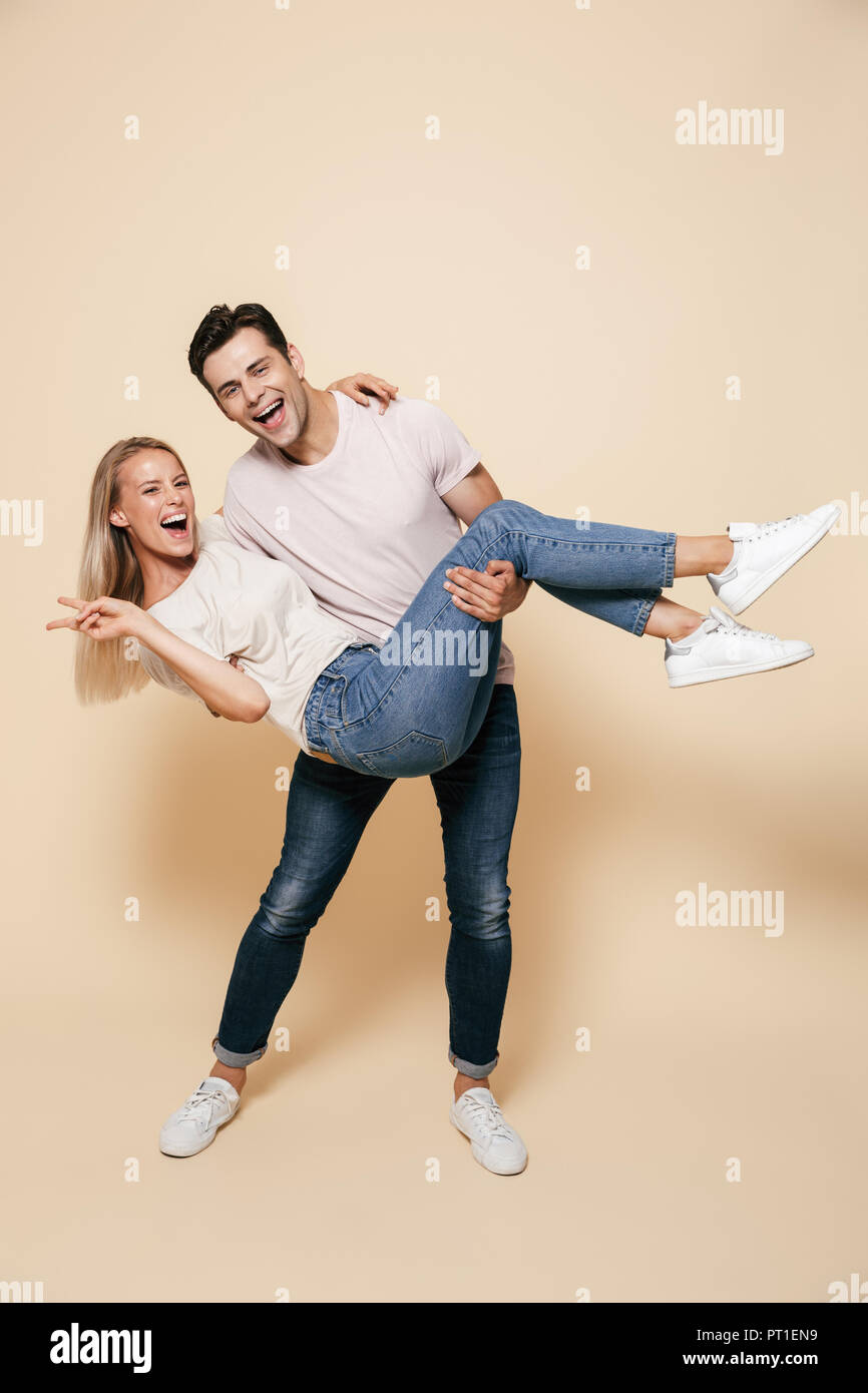 In voller Länge Porträt einer fröhlichen jungen Paar ständigen zusammen über beigen Hintergrund, Mann, Freundin auf seine Arme Stockfoto