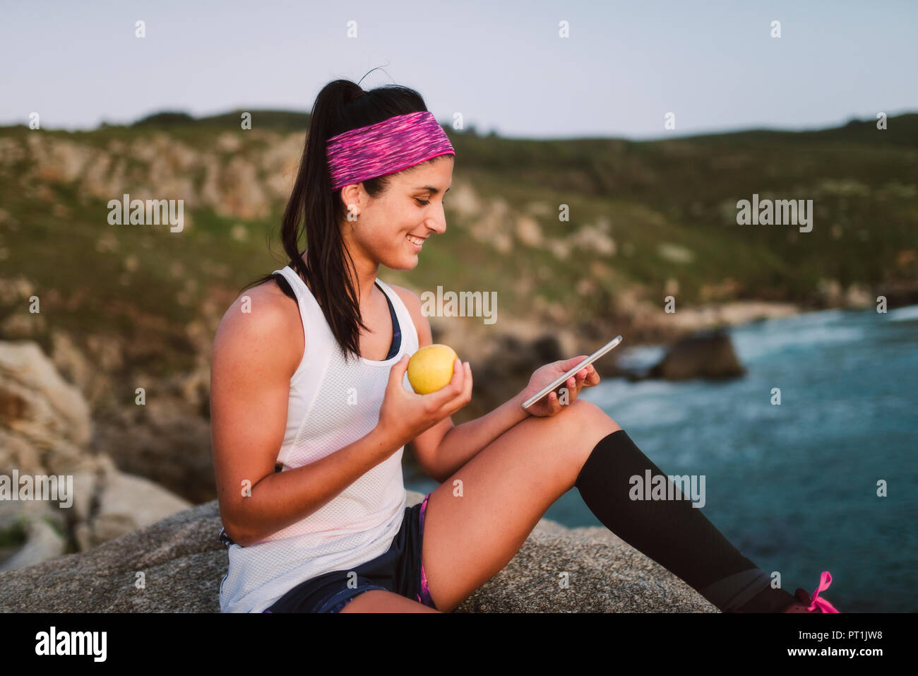 Sportliche Frau sitzen auf den Felsen am Abend, Apple und halten Sie Ihr Smartphone Stockfoto