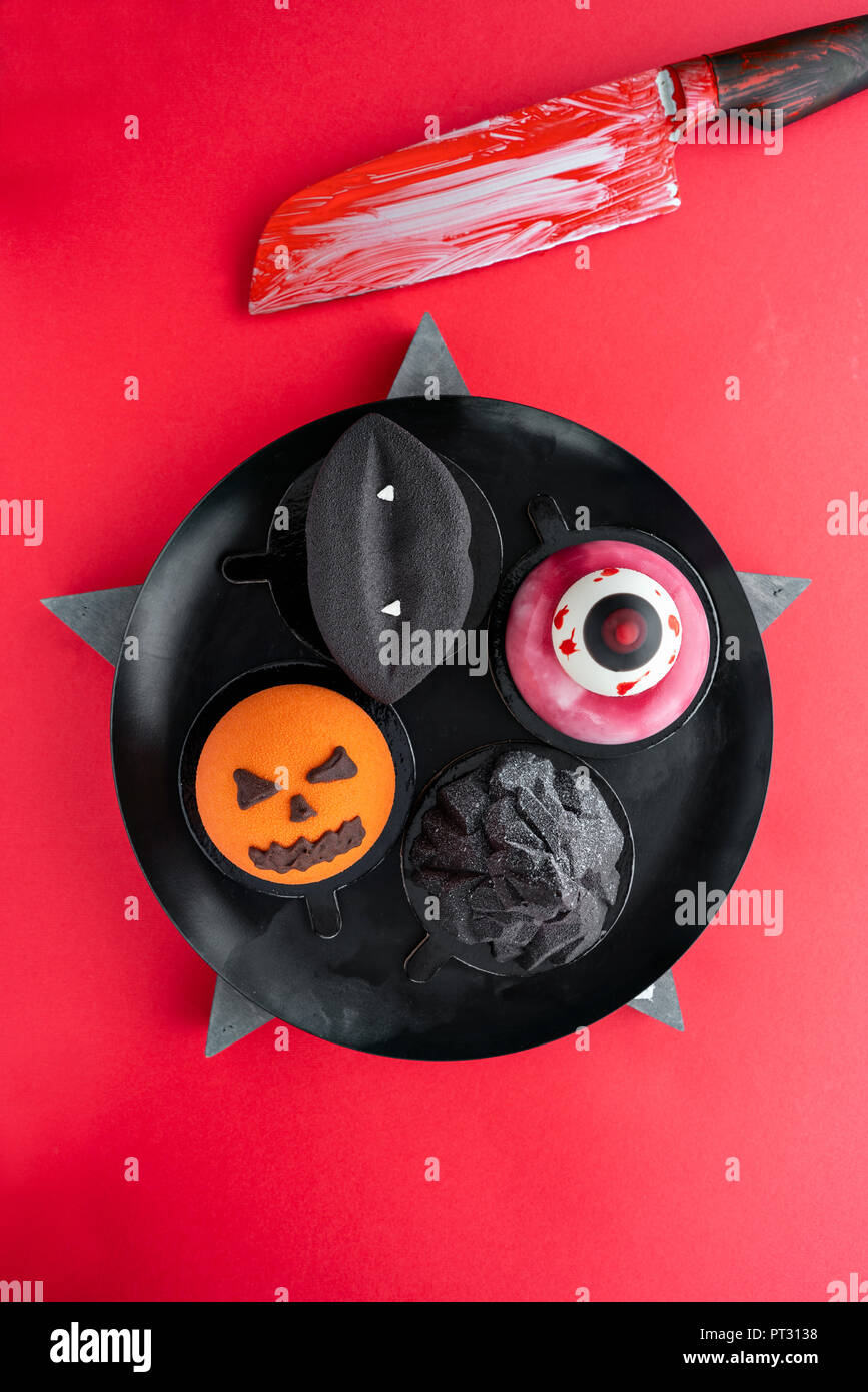 Schwarze Lippen mit Vampirzähnen, einer zerrissenen Auge, ein ominöser Kürbis und Black Mountain. Halloween scary Mousse Cake mit Kürbis auf rote Farbe Hintergrund, Ansicht von oben. Messer mit Blut Stockfoto