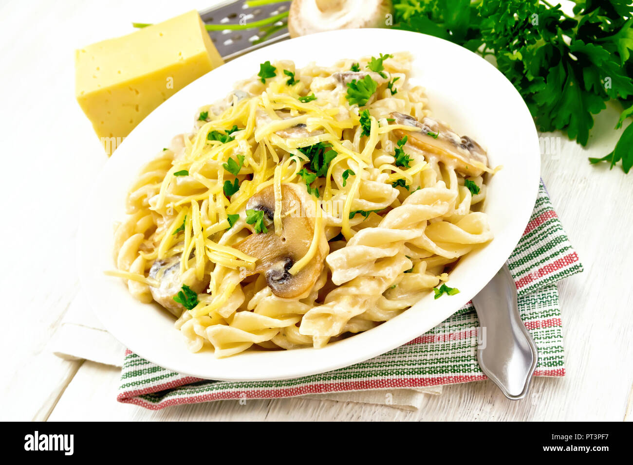 Fusilli mit Champignons in Rahmsauce, Petersilie und geriebenem Käse in einer Schüssel auf dem Handtuch gegen Licht Holzbrett Stockfoto