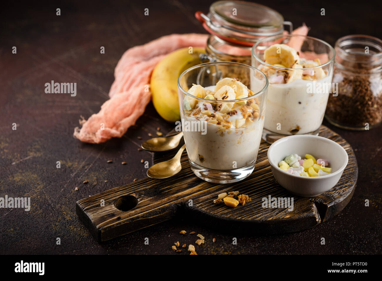 Gesundes Dessert aus Joghurt, eine Banane, Müsli und Nüsse auf einer hölzernen runde Schneidbrett auf einem dunklen Hintergrund Stockfoto