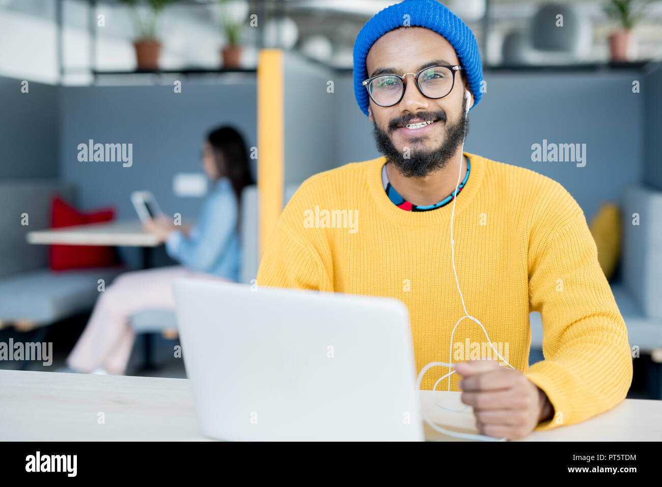 Gerne begeistert Junge arabische Kerl in Ohrhörer trägt blaue Strickmütze und gelben Pullover sitzen am Tisch und mit Laptop und Kamera suchen in Stockfoto