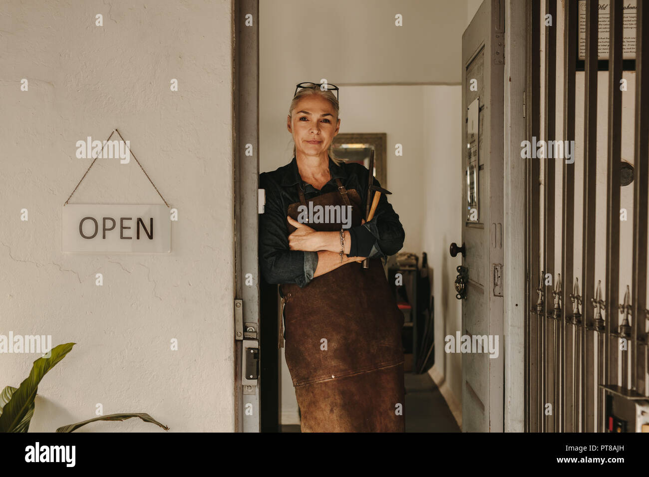 Portrait von älteren weiblichen Goldschmied stehend an Workshop Tür. Frau Juwelier tragen Schürze und Werkzeuge in der Werkstatt. Stockfoto