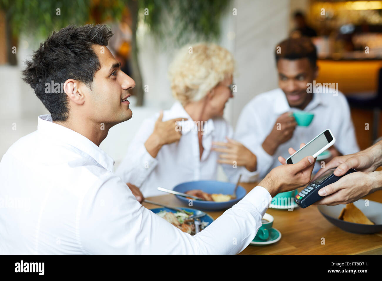 Gerne hübscher junger Mann im weißen Hemd am Tisch sitzen im Restaurant und die Kellnerin, während Sie ihr Smartphone für kontaktloses Bezahlen Stockfoto