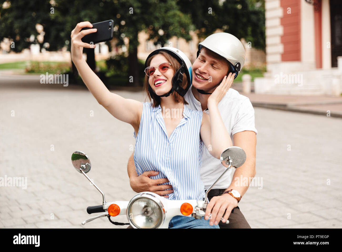 Glückliches junges Paar in Sturzhelme machen selfie auf dem Smartphone beim zusammen sitzen auf Roller im Freien Stockfoto
