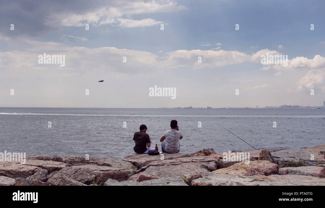 Anzeigen von zwei Jungen unkenntlich Männer sitzen auf den Felsen durch den Bosporus. Das Bild ist in der Gegend namens Moda befindet sich auf der asiatischen Seite von erfasst Stockfoto