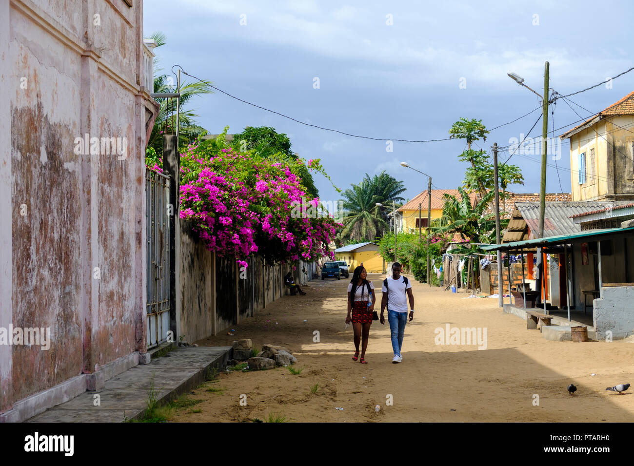 GRAND BASSAM, COTE DIVOIRE - 9. APRIL 2018: Junges Paar Spaziergang durch Bougainvillea gesäumten Straße im alten kolonialen Stadtteil der ehemaligen Hauptstadt von Côte d'Ivoire Stockfoto