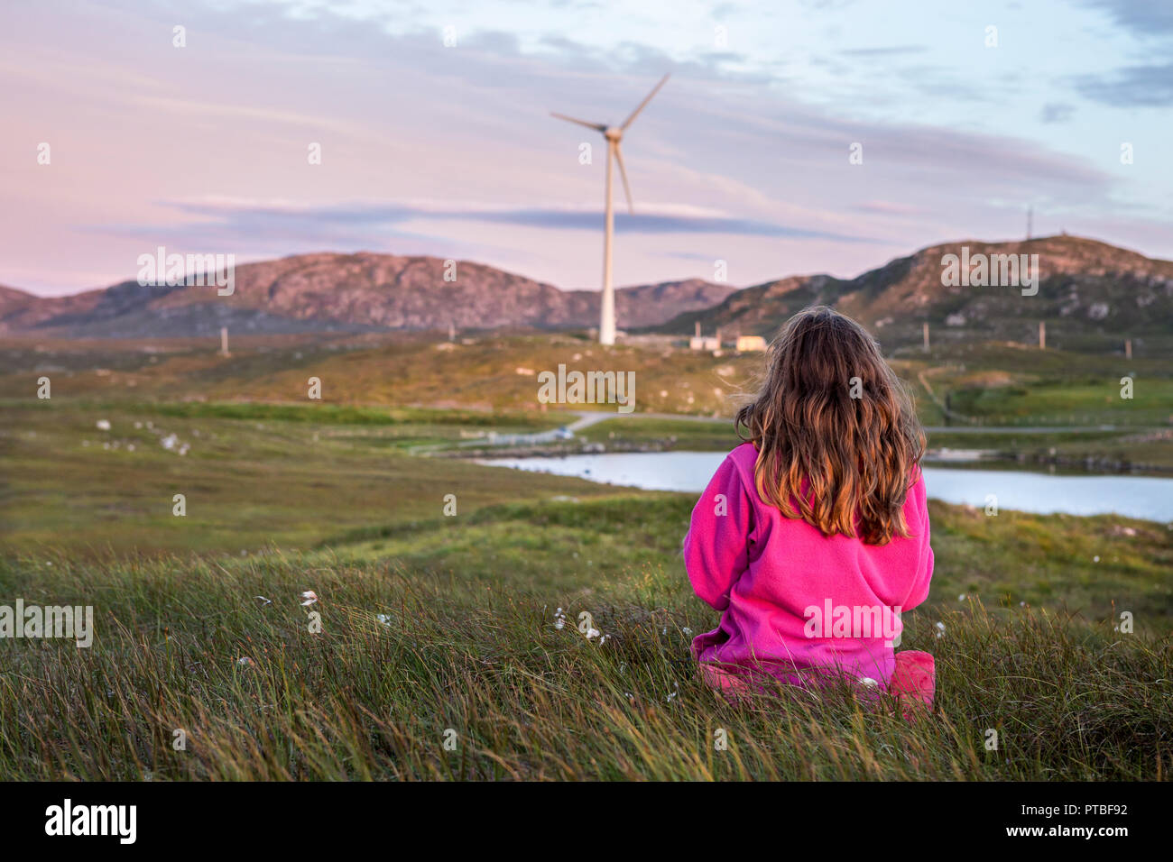 9 Jahre altes Mädchen von hinten gesehen, das in einem sitzt Wiese in schottischer Landschaft bei Sonnenuntergang mit Windturbine in Die Entfernung Stockfoto