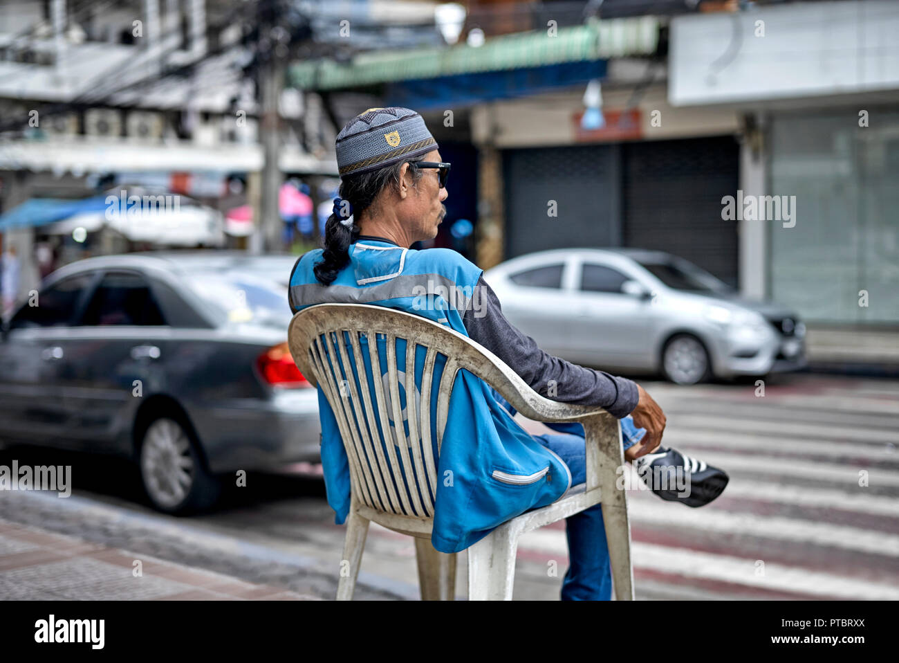 Mann, der draußen auf einem Plastikstuhl auf einer thailändischen Straße sitzt und die Tageszeit vergeht. Südostasien, Stockfoto