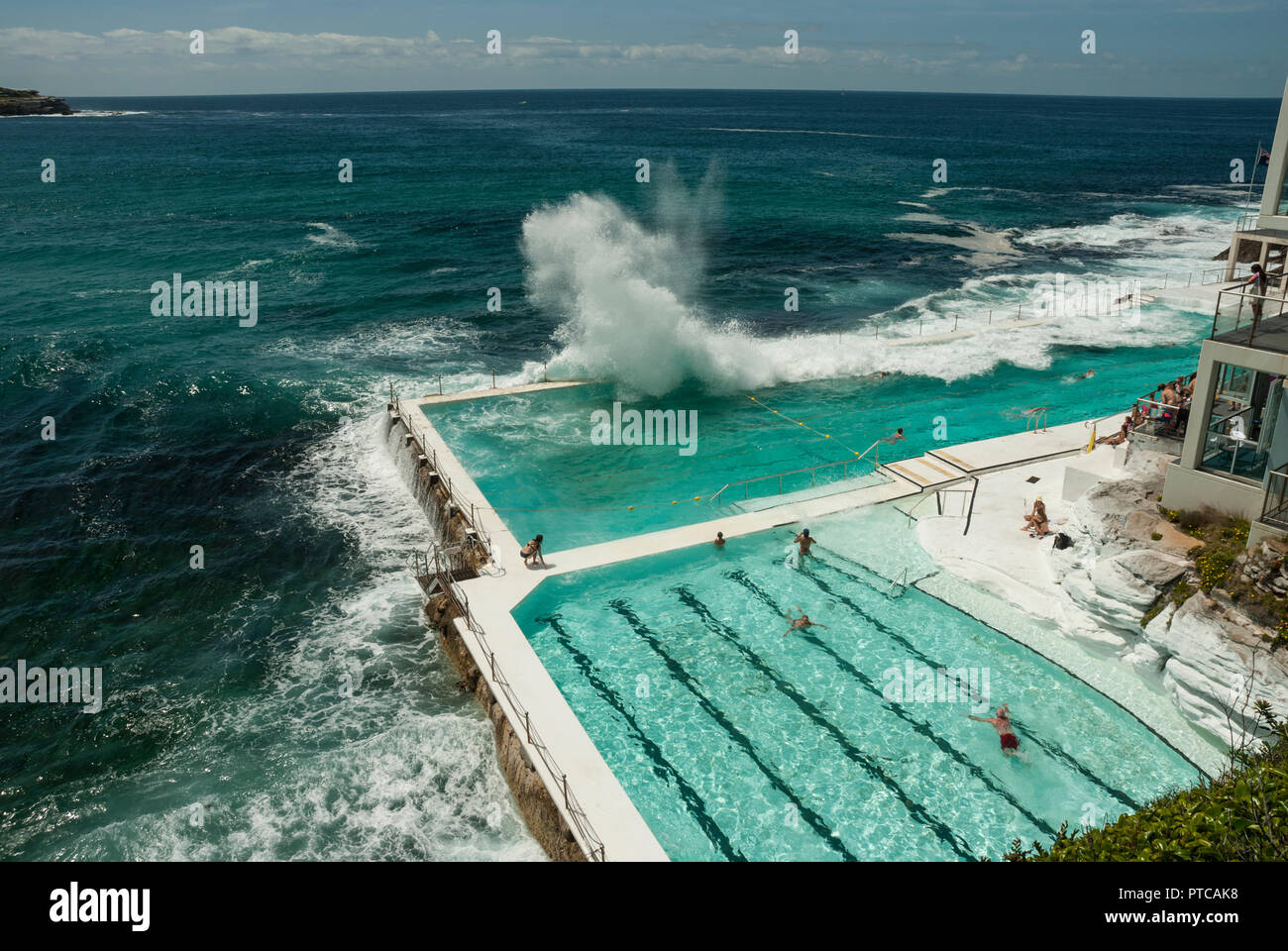 Riesige Wellen über Bondi Iceberg Meerwasser Außenpools (Sydney). Sonnenschein und attraktive Blau Grün Pools mit einige Leute schwimmen. Stockfoto