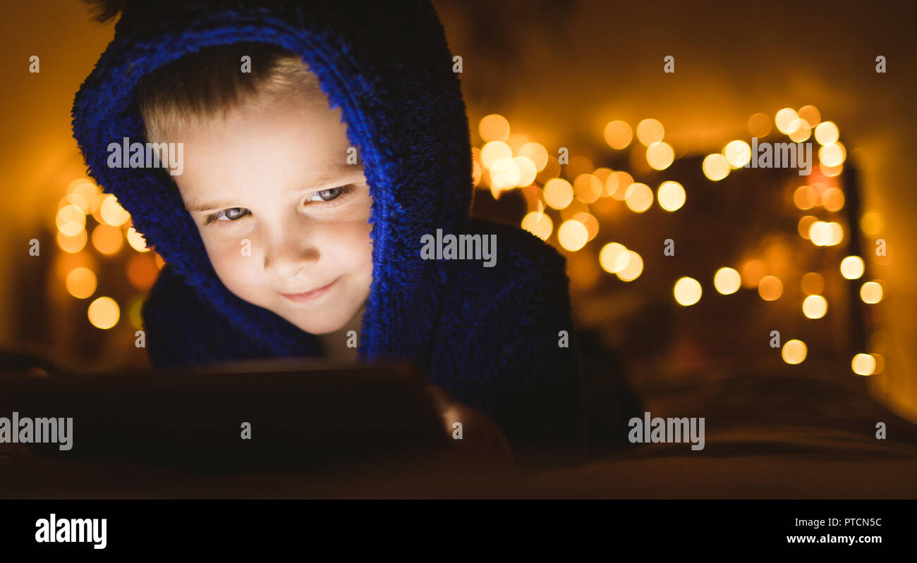 Junge in der blauen Jacke mit digitalen Tablette gegen Weihnachtsbeleuchtung Stockfoto