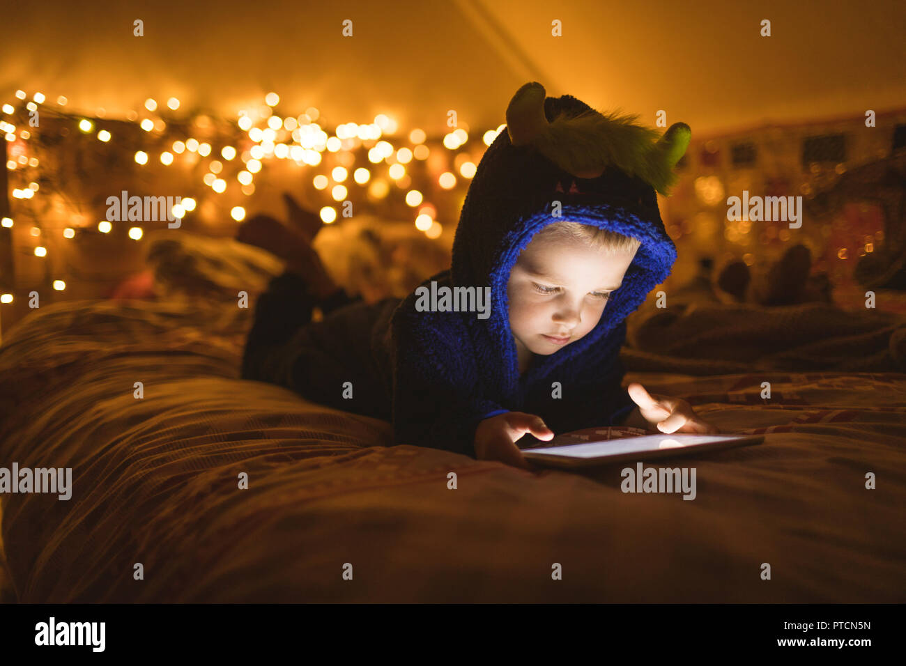 Junge in der blauen Jacke mit digitalen Tablette gegen Weihnachtsbeleuchtung Stockfoto