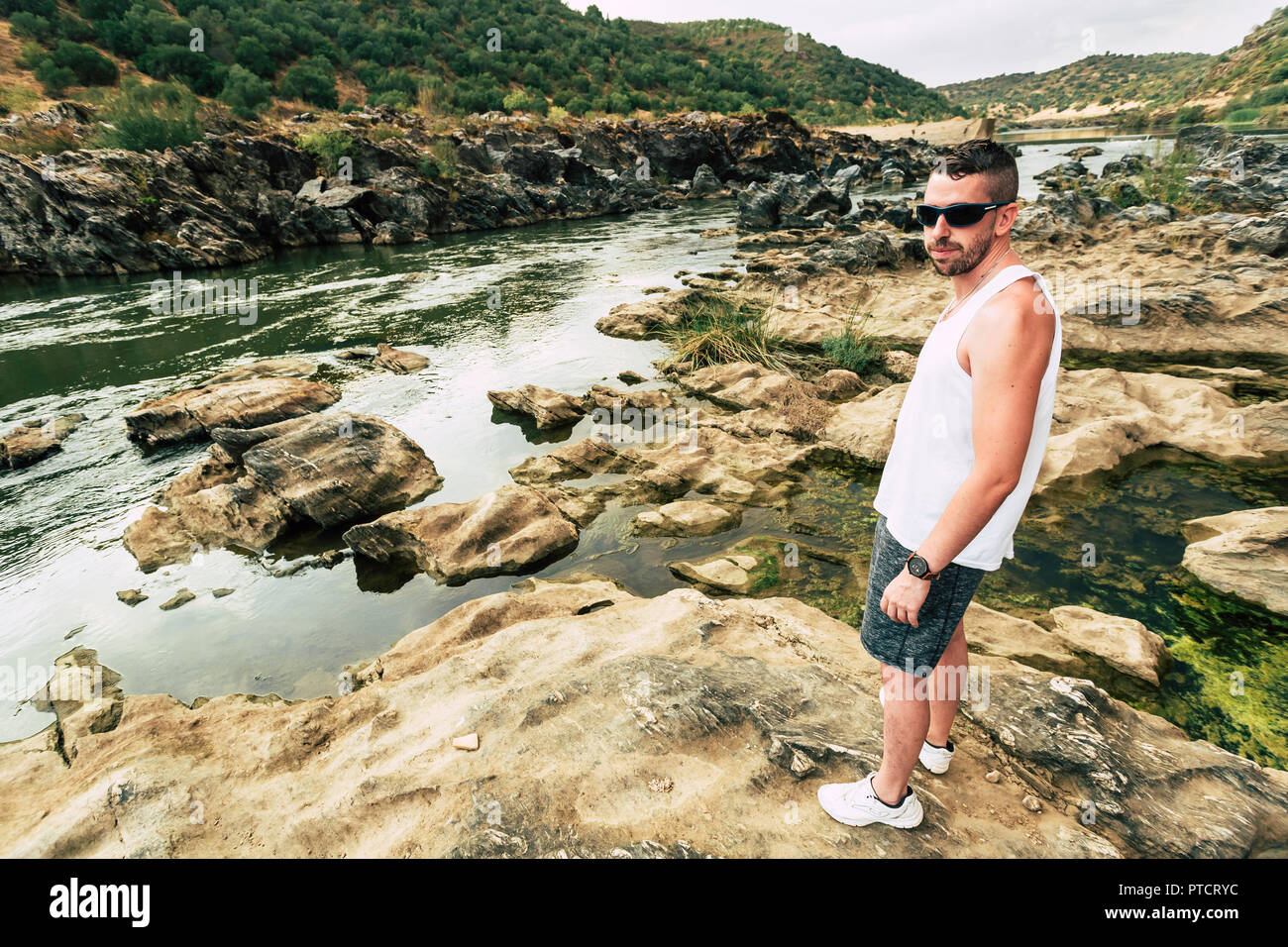 Junger Mann mit Bart, der auf den Felsen des Flusses Guadiana mit den Bergen im Hintergrund Stockfoto