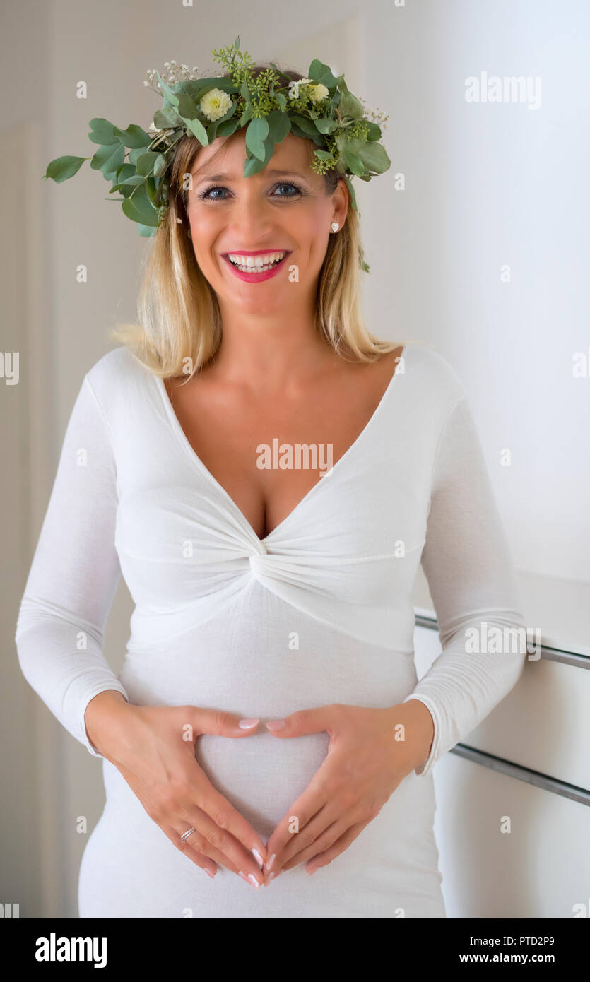 Frau, Braut, mit Kranz aus Blumen, neun Monate schwanger, mit den Händen bilden ein Herz vor Ihren schwangeren Bauch, Deutschland Stockfoto