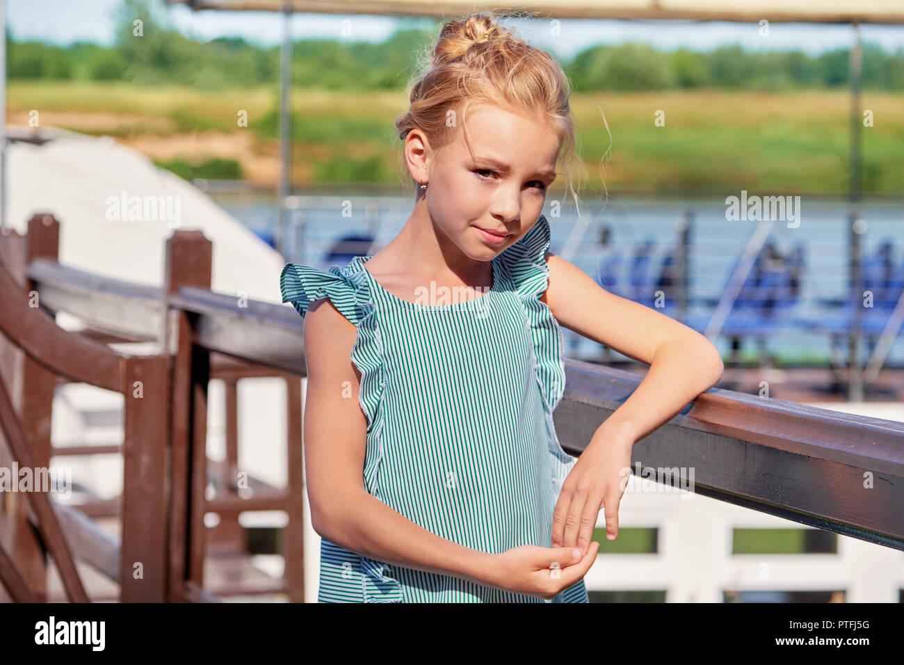 Portrait elegante junge Mädchen von 9-11 Jahre im Freien, ihre Ellenbogen stützte sich auf das Geländer. Niedliche Kind blond lächelnd, sonnigen Sommertag auf der Pier. Fashion kid Konzept. gestreiftes Kleid, maritimen Stil. Stockfoto