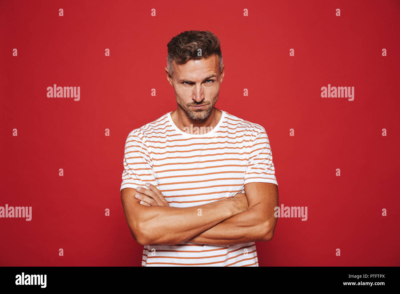 Mann 30 s in gestreiften T-Shirt stand mit verschränkten Armen und wütenden Blick über den roten Hintergrund irritiert Stockfoto