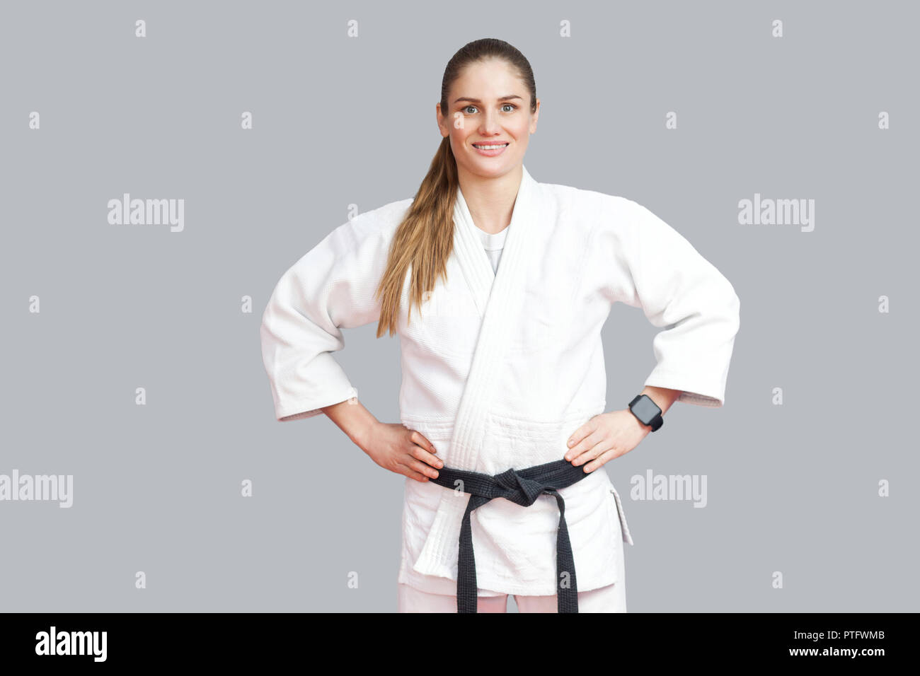 Gerne fröhliche schöne blonde Junge athletische Frau in weißen Kimono, Schwarze Riemen stehen, holdind Hände an der Hüfte, toothy Lächeln und Kameras Stockfoto