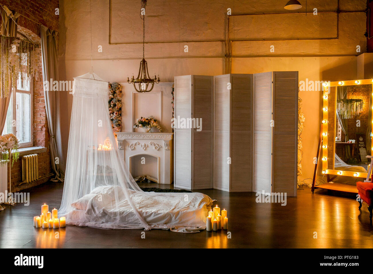 Loft-stil Zimmer mit einem Bett, einem Baldachin, ein weißer Kamin mit einem Blumenarrangement, einen weißen Bildschirm, einem großen Spiegel und brennende Kerzen Stockfoto