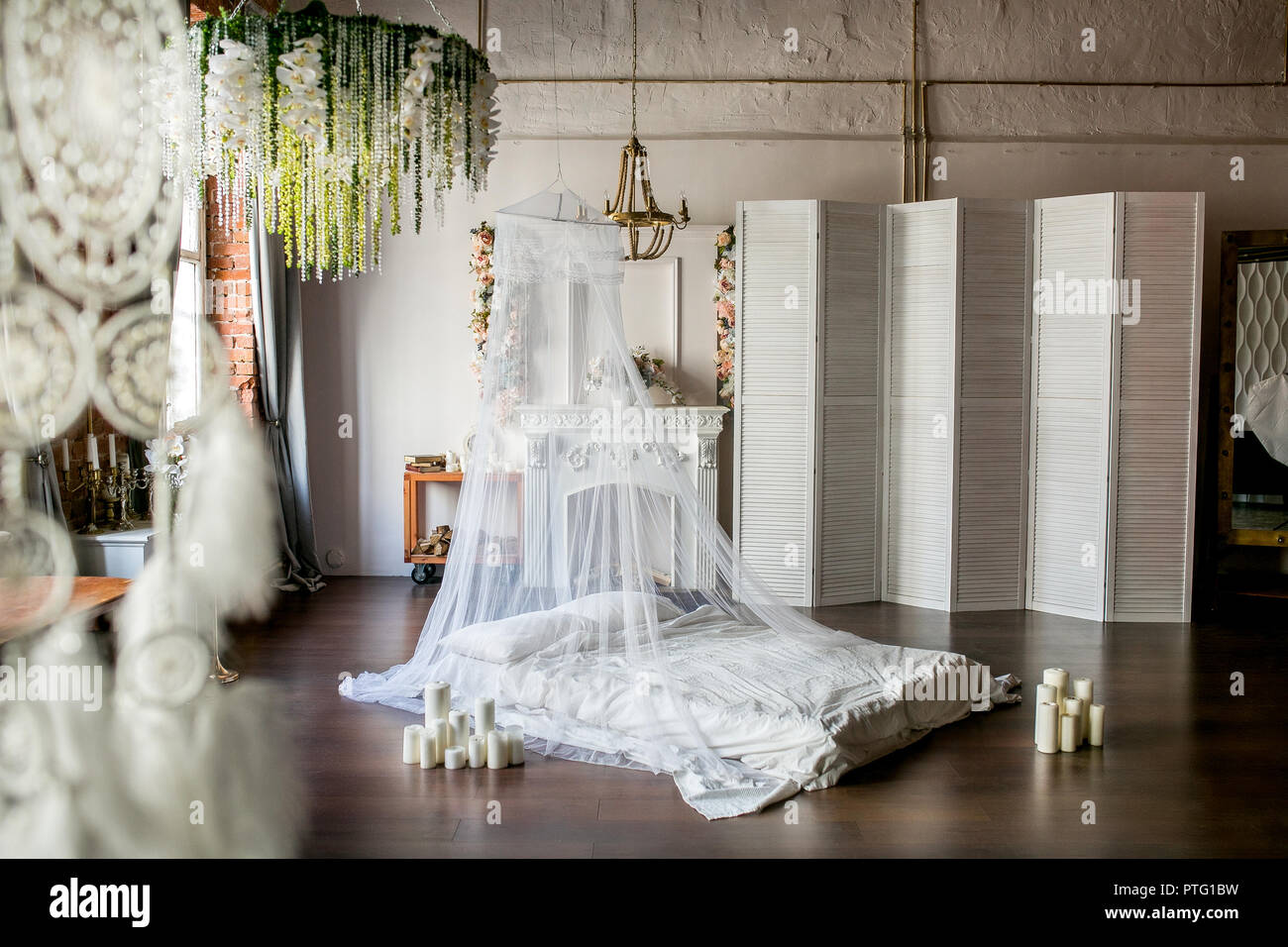 Loft-stil Zimmer mit einem Bett, einem Baldachin, ein weißer Kamin mit einem Blumenarrangement, einen weißen Bildschirm, einem großen Spiegel und Kerzen Stockfoto