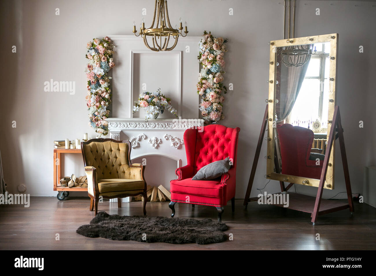 Helle Loft Zimmer mit einem roten Sessel, einen braunen Sessel, ein weißer Kamin mit Blumen, einem großen Spiegel mit Lampen und einem Kronleuchter Stockfoto