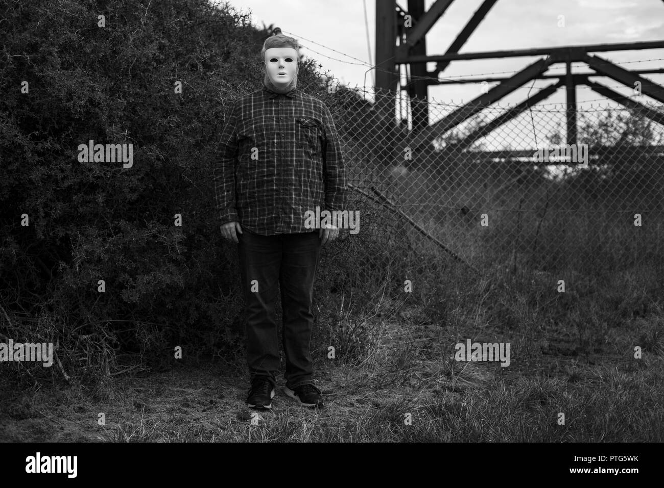 Creepy scary Mann in einer weißen Maske steht drohend Wartezeiten vor einem Busch und Zaun Stockfoto