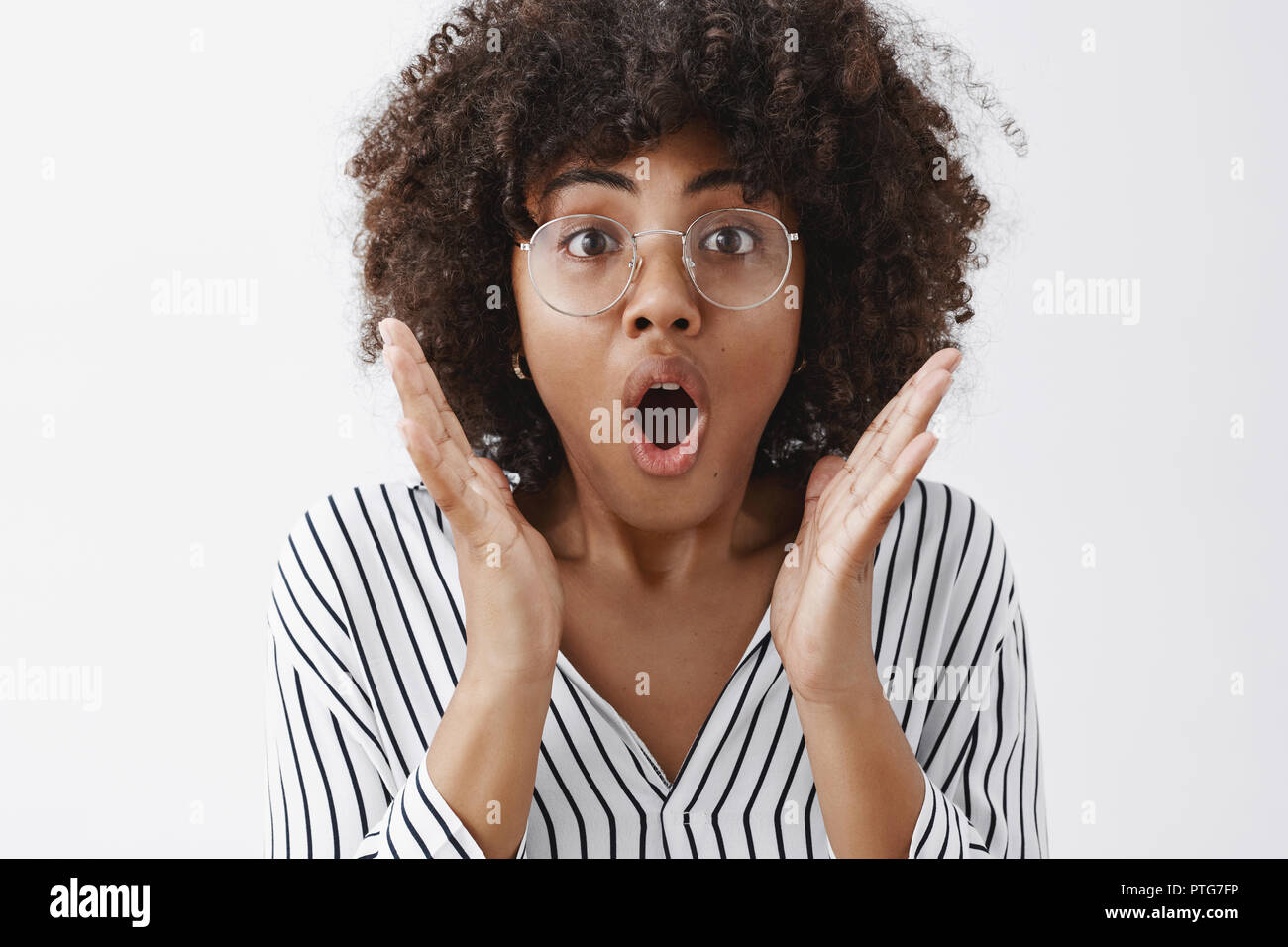 Headshot beeindruckt attraktive african american female mit Afro Frisur in transparenten Gläser keuchend öffnung öffnung von Staunen und Schock, Hände halten in der Nähe von Gesicht, schüttelte über graue Wand Stockfoto