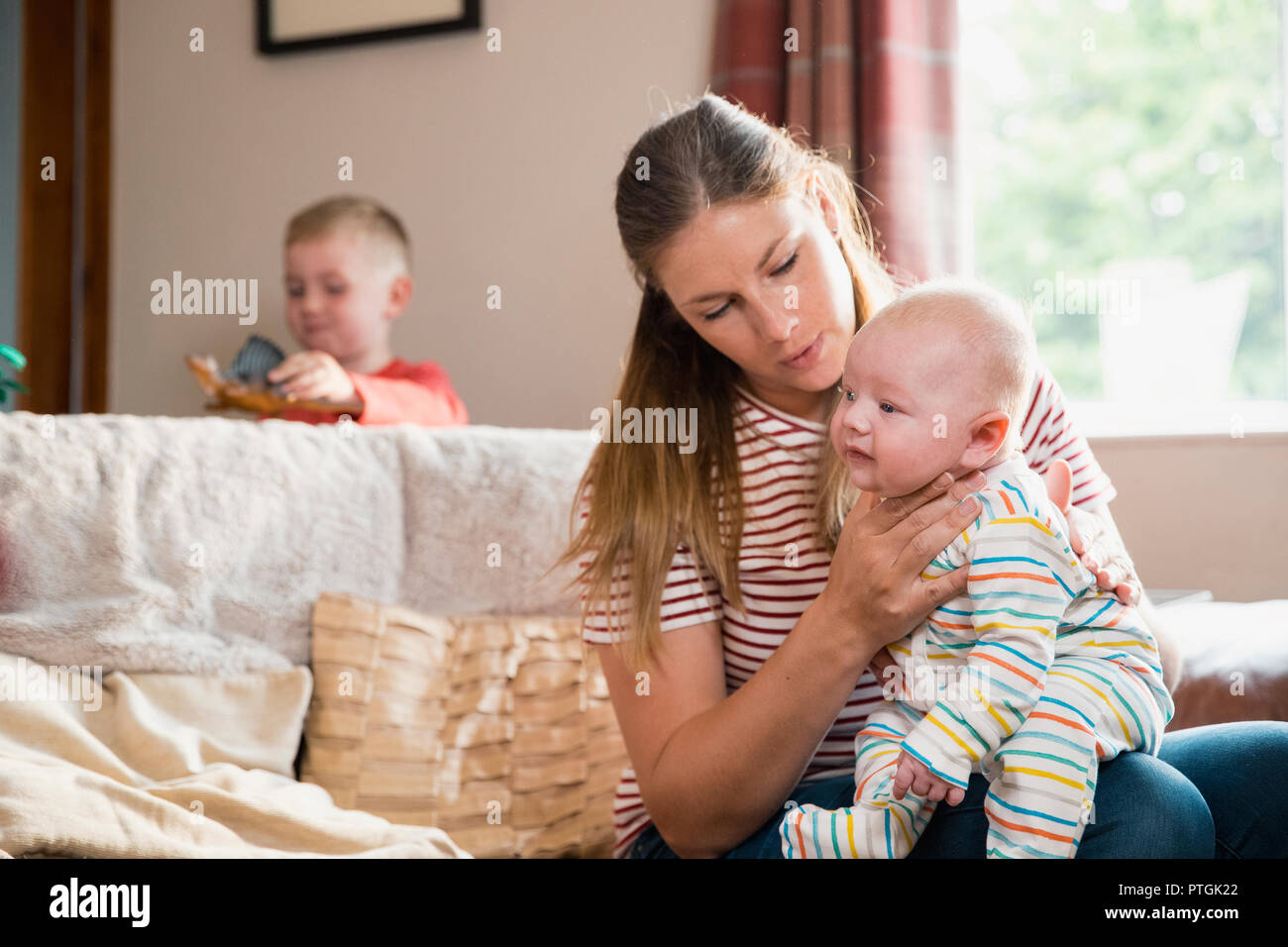 Mutter sitzt auf der Kante des Sofas im Wohnzimmer und Aufstoßen ihr neugeborenes Kind nach ihm zuführen. Ihr Sohn ist stadning hinter dem Sofa p Stockfoto