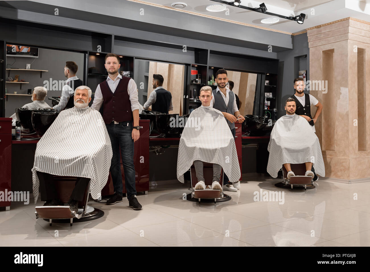 Drei professionelle Friseure stehen in der Nähe von Kunden in Barbershop.  Männliche Klienten sitzen in Friseur Haarschnitt Stühle mit gestreiften  Gewändern bedeckt. Männer posieren und Kamera Stockfotografie - Alamy