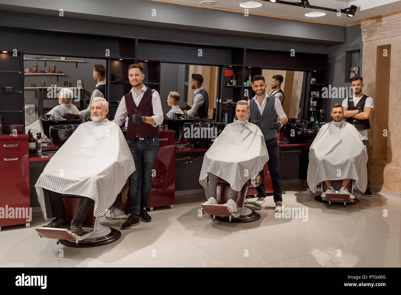 Männliche Klienten sitzen in Friseur Haarschnitt Stühle mit gestreiften Gewändern bedeckt. Drei professionelle Friseure stehen in der Nähe von Kunden in Barbershop. Männer posieren und Kamera. Stockfoto