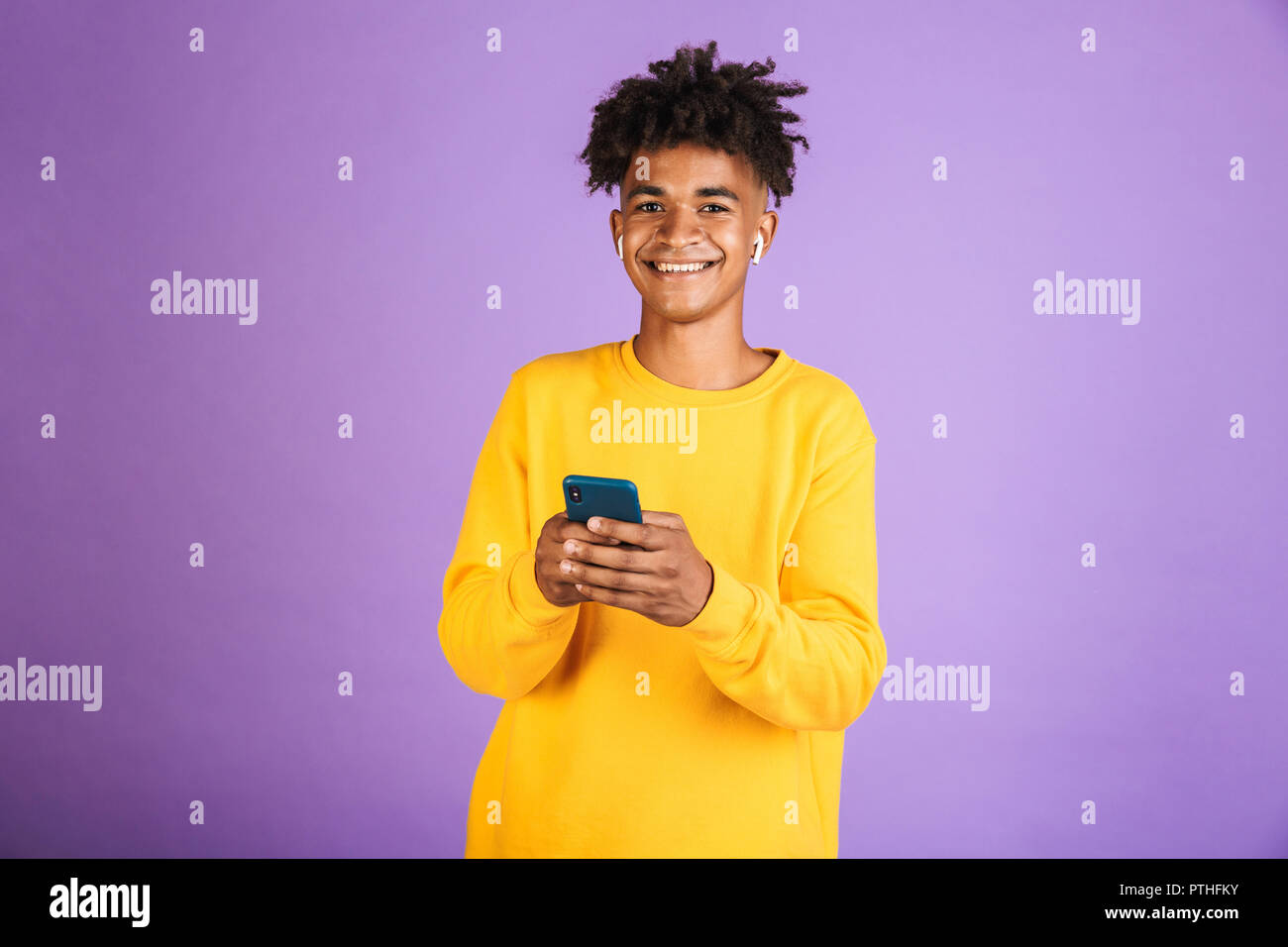 Portrait von jugendlich afrikanische amerikanische Mann und lächelnd Smartphone, bluetooth Kopfhörer über violett Hintergrund isoliert Stockfoto