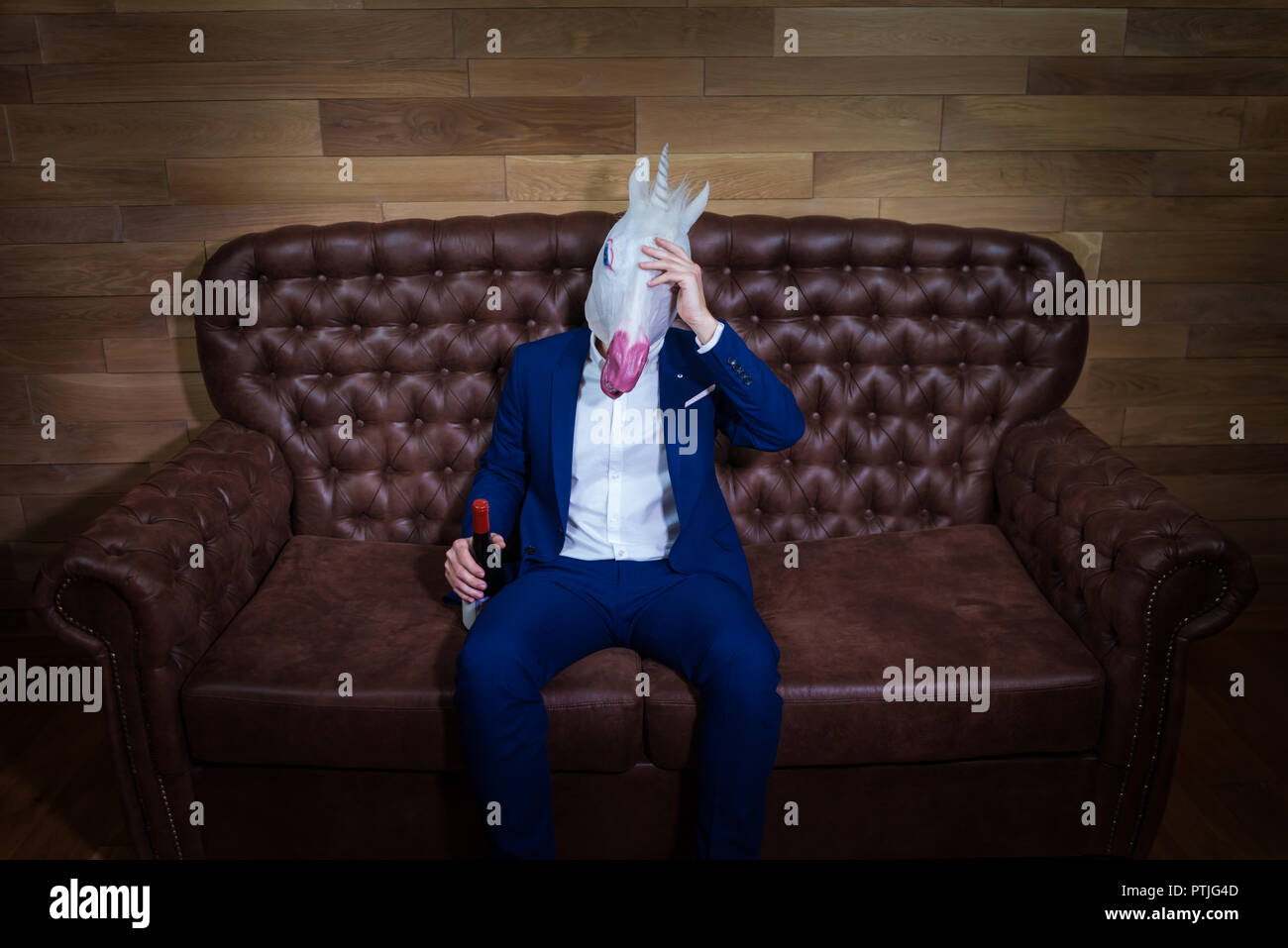 Lustige Einhorn im eleganten Anzug sitzt auf einem Sofa mit einer Flasche Wein. Ungewöhnliche Boss mit Kopfschmerzen zu Hause. Freaky Mann in komischen Maske. Stockfoto