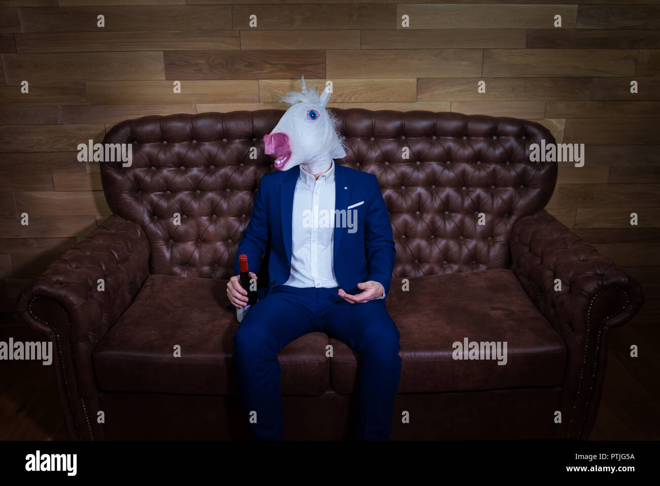 Lustige Einhorn im eleganten Anzug sitzt auf einem Sofa mit einer Flasche Wein. Ungewöhnliche Mann mit Kopfschmerzen zu Hause. Freaky Chef in komischen Maske. Stockfoto