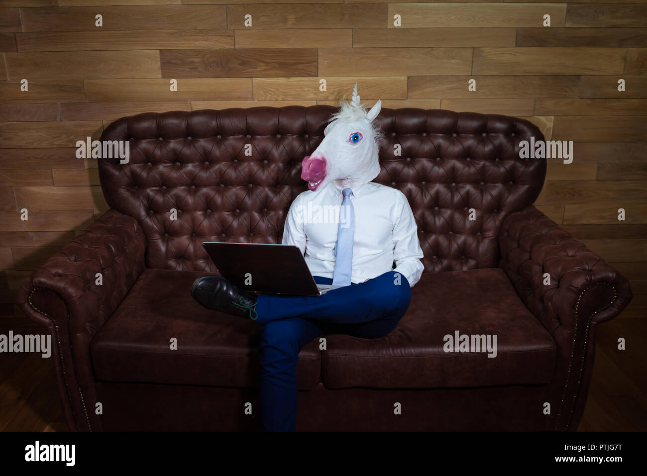 Stilvolle Mann im eleganten Anzug zu Hause arbeiten im Büro. Ungewöhnliche junge Manager in komischen Maske auf dem Hintergrund der hölzernen Wand. Lustige Einhorn sitzt auf einem Sofa. Stockfoto