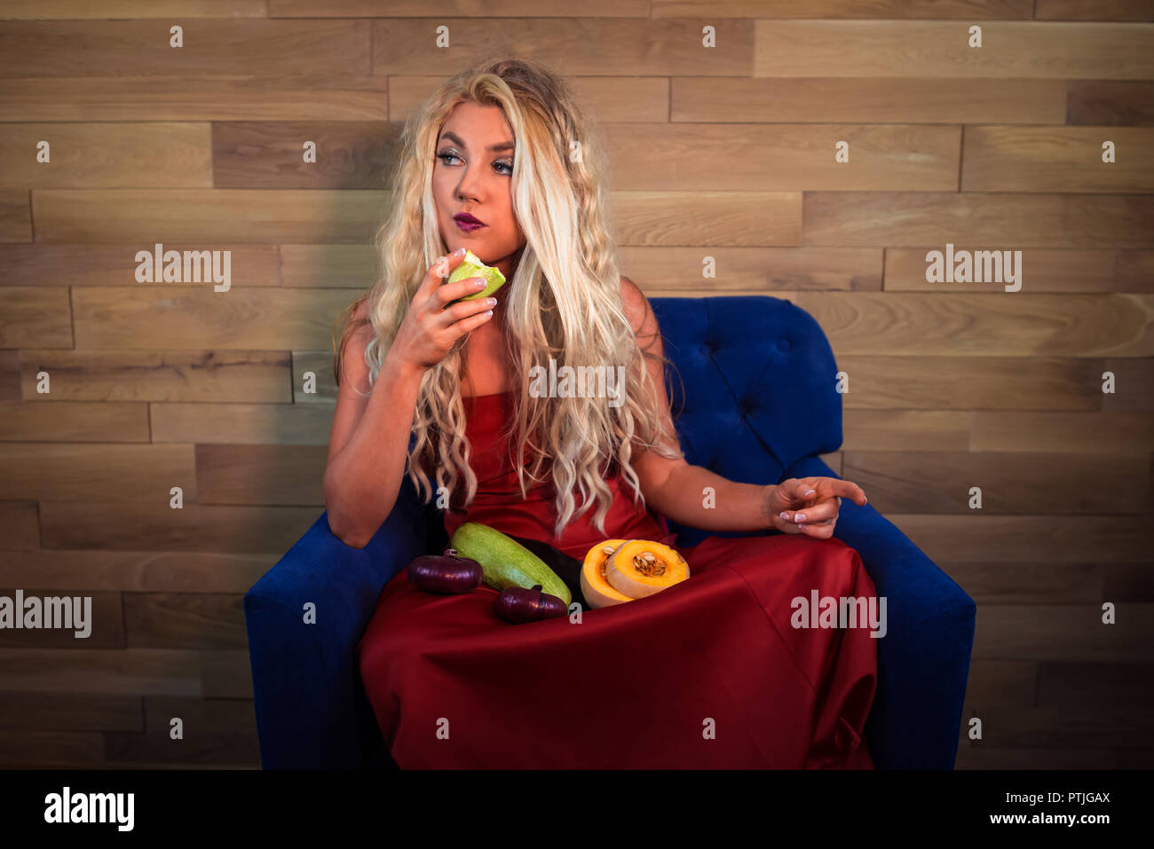 Junge vegetarische Frau im eleganten roten Kleid sitzt auf einem Sessel auf den Hintergrund der hölzernen Wand und hält Essen in die Hände. Unglücklich Hausfrau isst Gemüse. Stockfoto